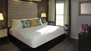 Una cama grande con cabecera, dos mesitas de noche con lámparas, alfombra estampada y un florero con flores sobre un tocador.
