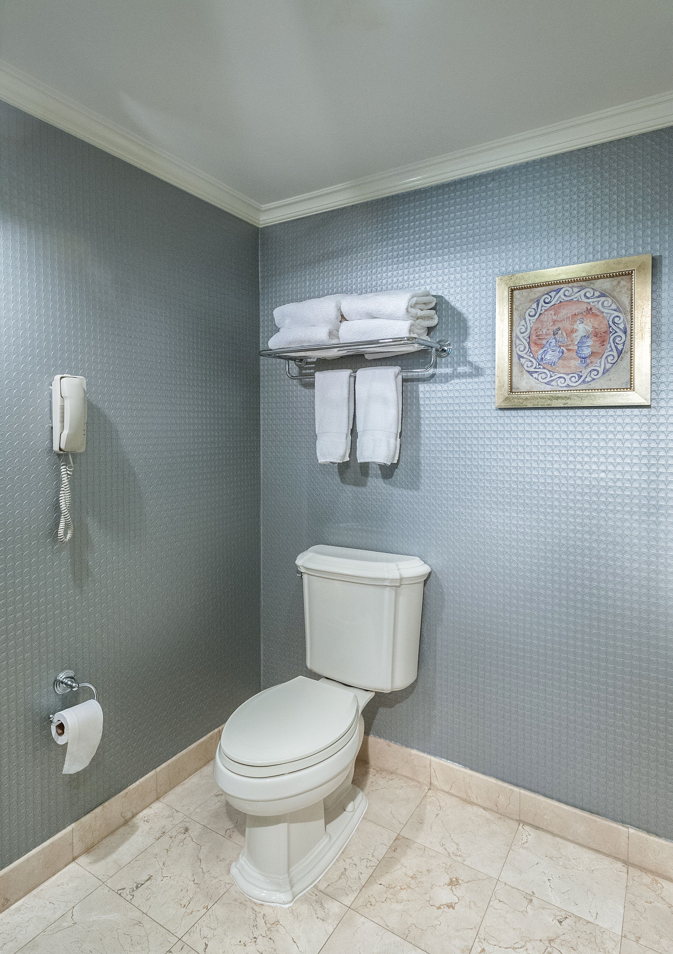 A bathroom with a toilet, a towel rack with towels, a framed picture on the wall, a telephone, and a toilet paper holder.