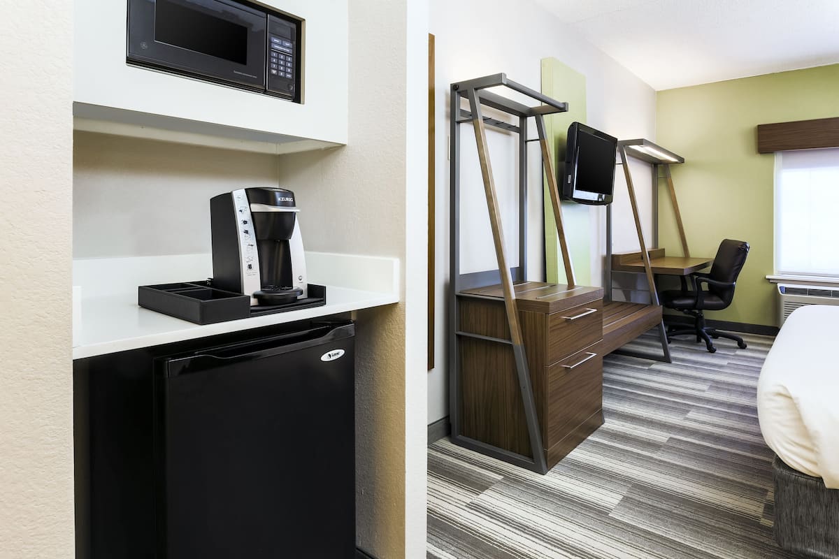 Une chambre d’hôtel compacte comprenant un coin cuisine, un lit, un bureau avec une chaise et un téléviseur fixé sur un support en bois.
