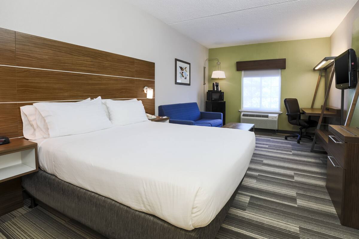Une chambre d’hôtel avec un grand lit, un bureau avec une chaise, une télévision et une fenêtre avec des stores.