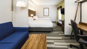 A hotel room with a blue sofa, a wooden coffee table, a bed with white bedding, a desk with a chair, and a TV mounted on the wall.