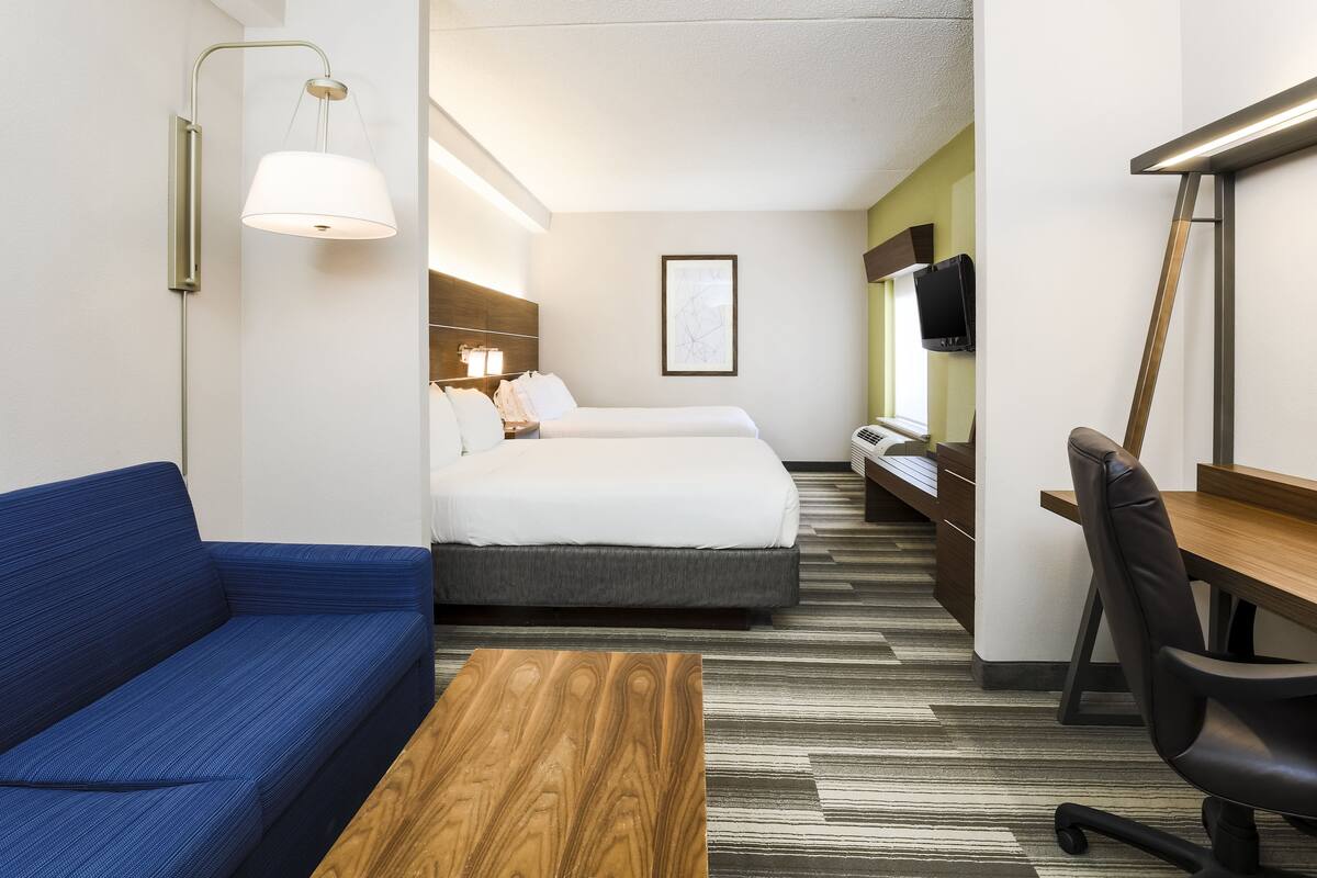 Une chambre d’hôtel avec un canapé bleu, une table basse en bois, un lit avec une literie blanche, un bureau avec une chaise et un téléviseur fixé au mur.