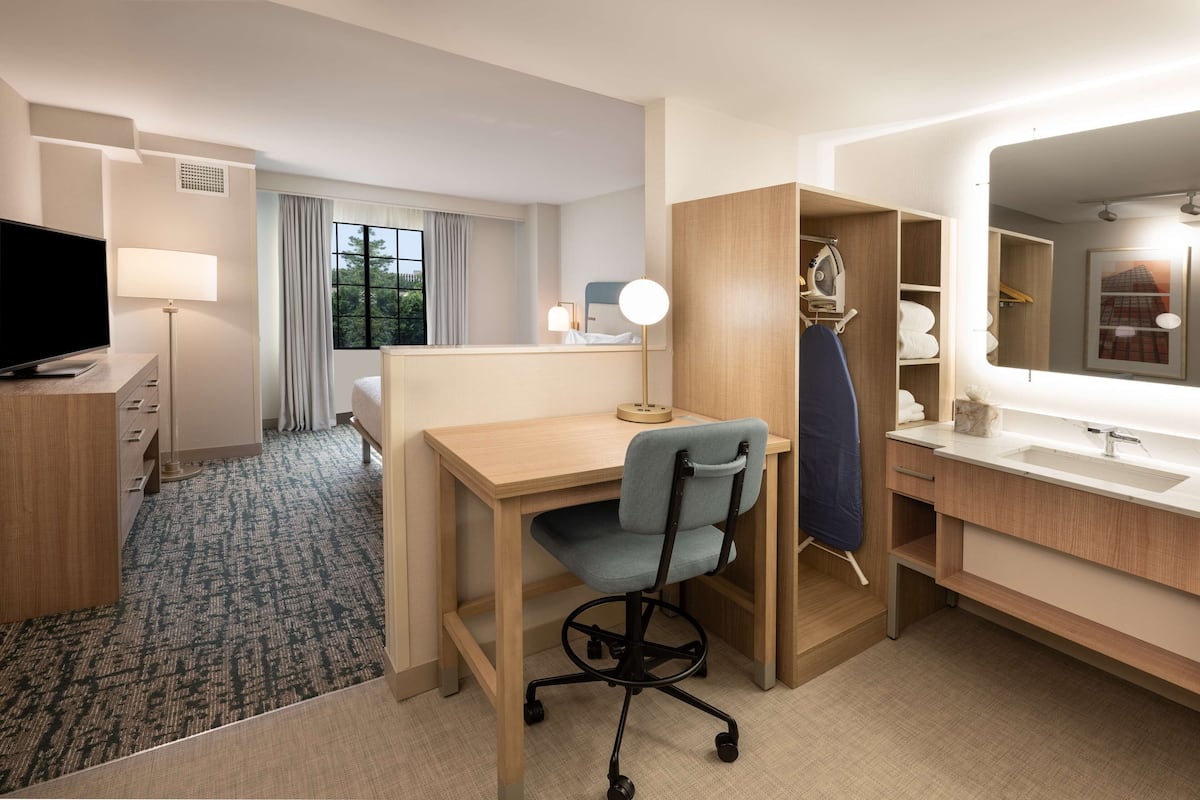 Hotellihuone, jossa on työpöytä, tuoli, televisio sekä kylpyhuone, jossa on peili ja pesuallas.