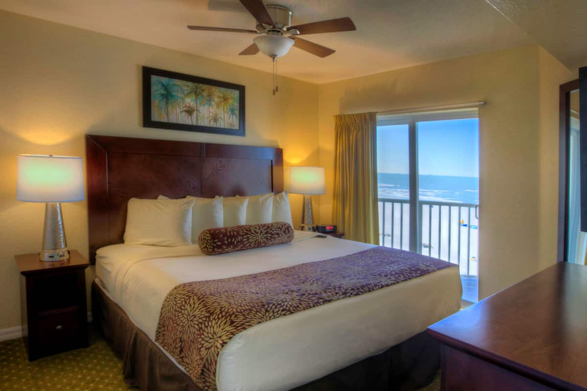 A hotel room with a large bed, two bedside lamps, a ceiling fan, a painting on the wall, and a view of the beach through the window.