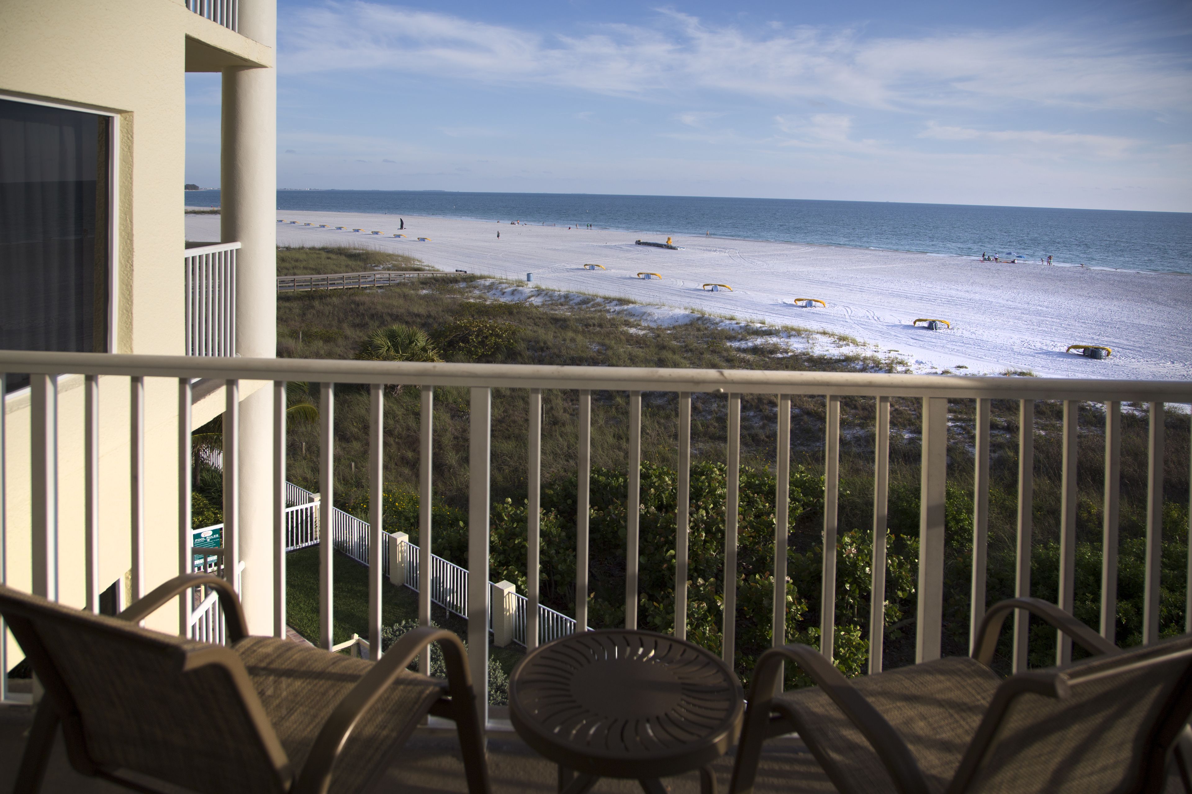 A balcony with chairs and a table overlooking a beach with umbrellas.