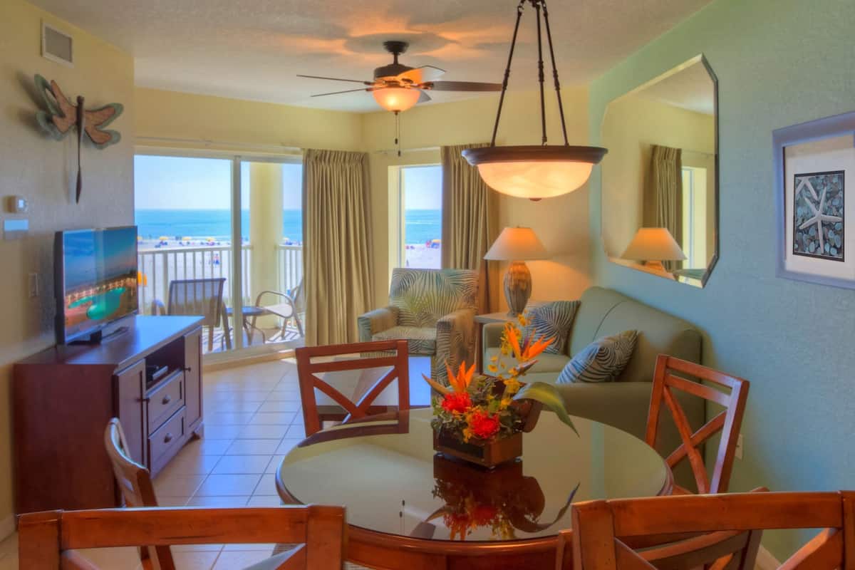 A room with a dining table, chairs, a TV, a ceiling fan, and a view of the ocean through large windows.