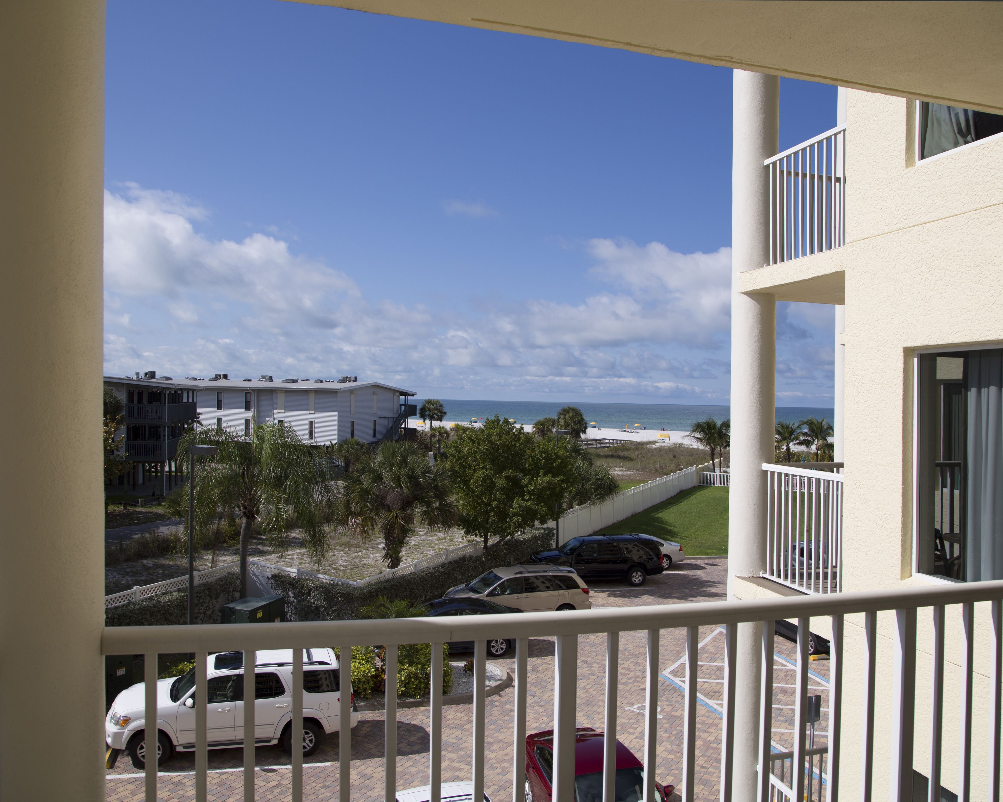 A balcony with a view of a beach, parked cars, and a building with a white exterior.