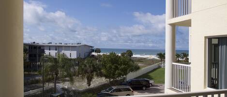 A balcony with a view of a beach, parked cars, and a building with a white exterior.