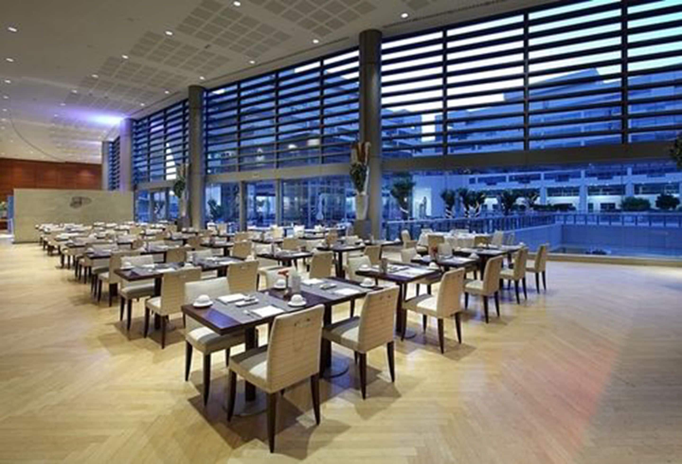 Un vaste espace dédié à la salle à manger, avec de nombreuses tables et chaises, de grandes fenêtres équipées de stores horizontaux et une vue sur le paysage urbain.