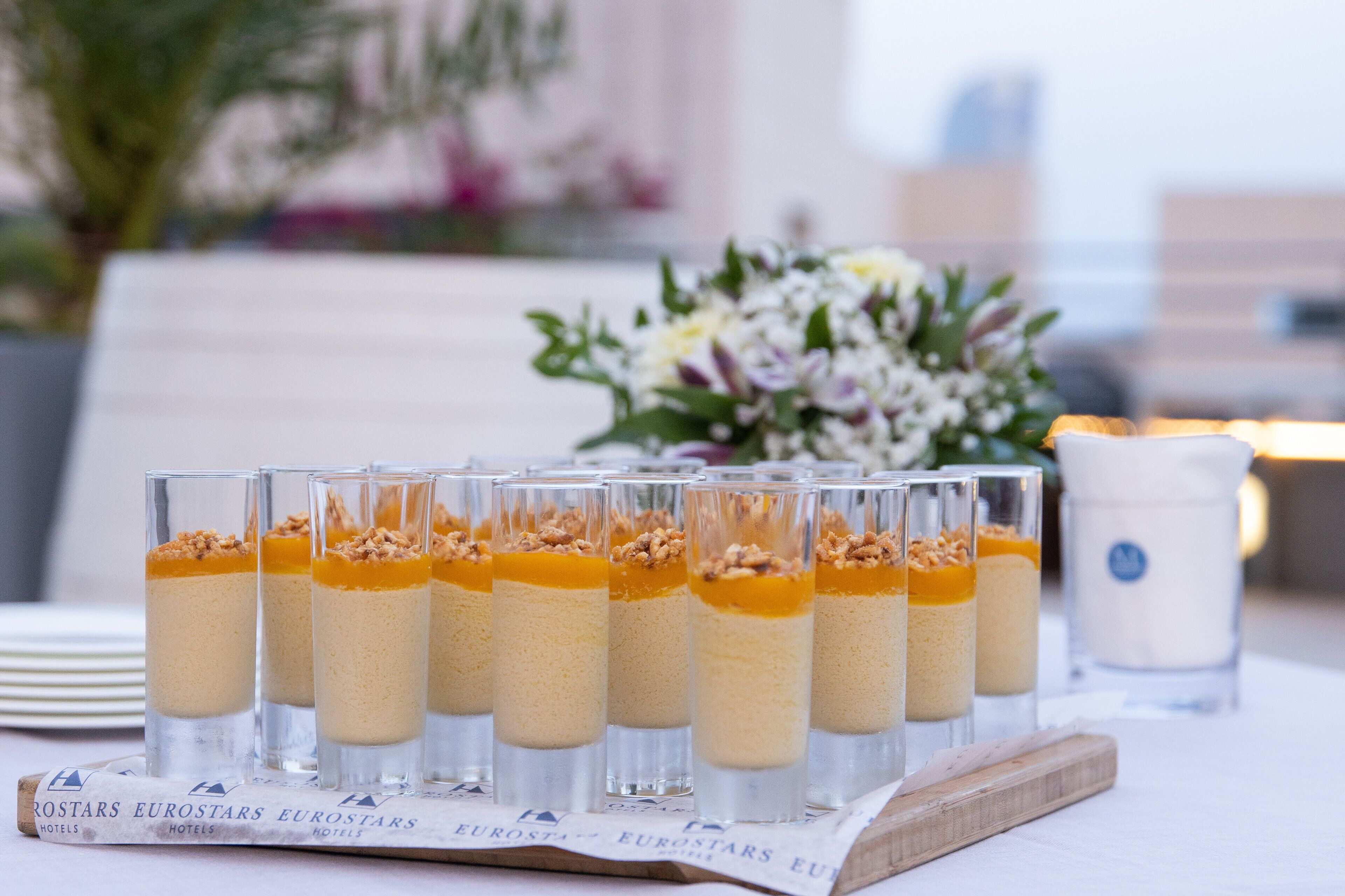 A table with dessert glasses arranged on a wooden board, a candle, and a bouquet of flowers in the background.