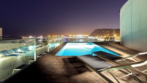 Una piscina en la azotea con sillones de descanso y vistas a la ciudad de noche.