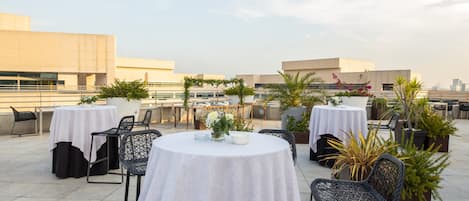 Una terraza en la azotea con mesas y sillas redondas, rodeada de plantas y flores en macetas.
