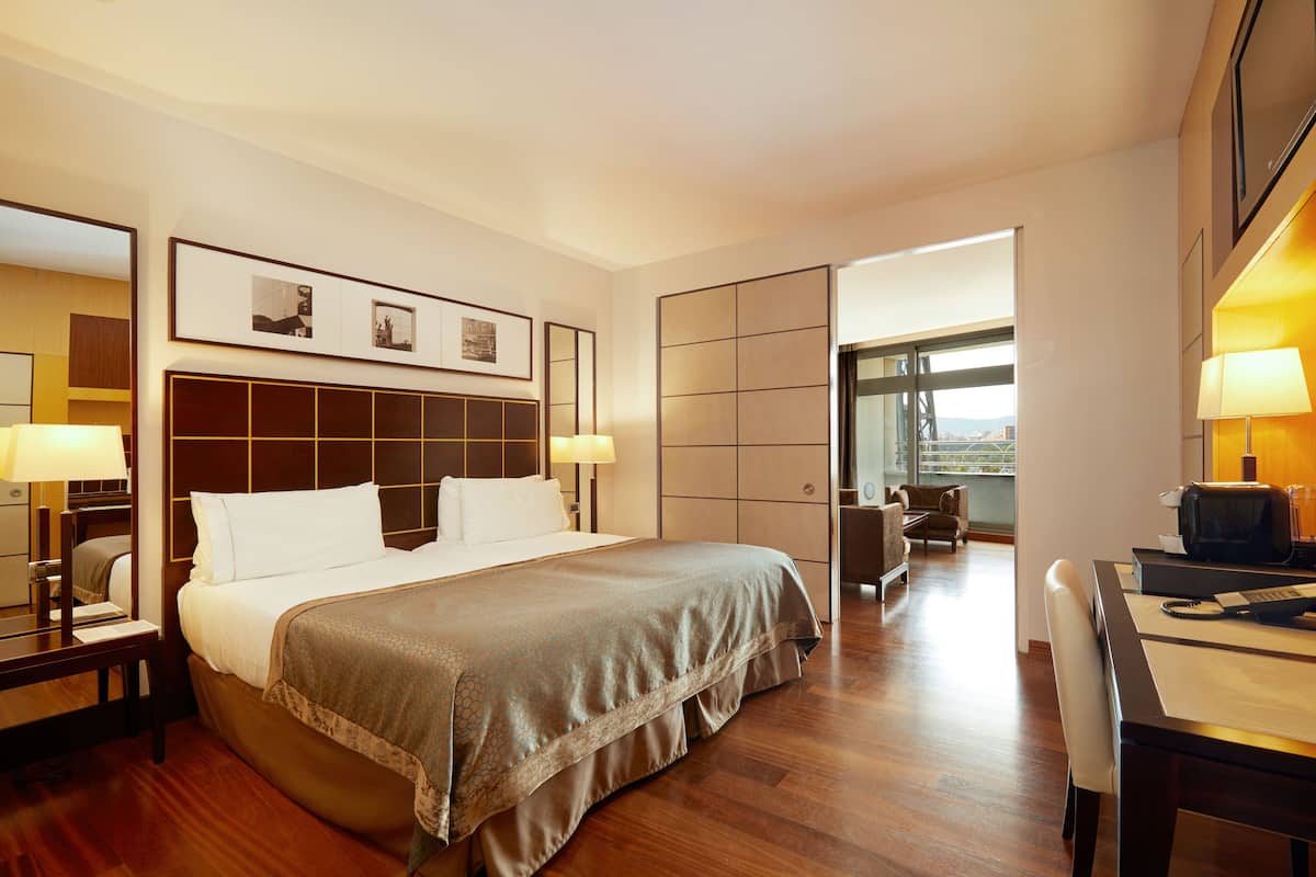 Een hotelkamer met een groot bed, nachtkastjes met lampen, een bureau met stoel, en uitzicht door een open deur.