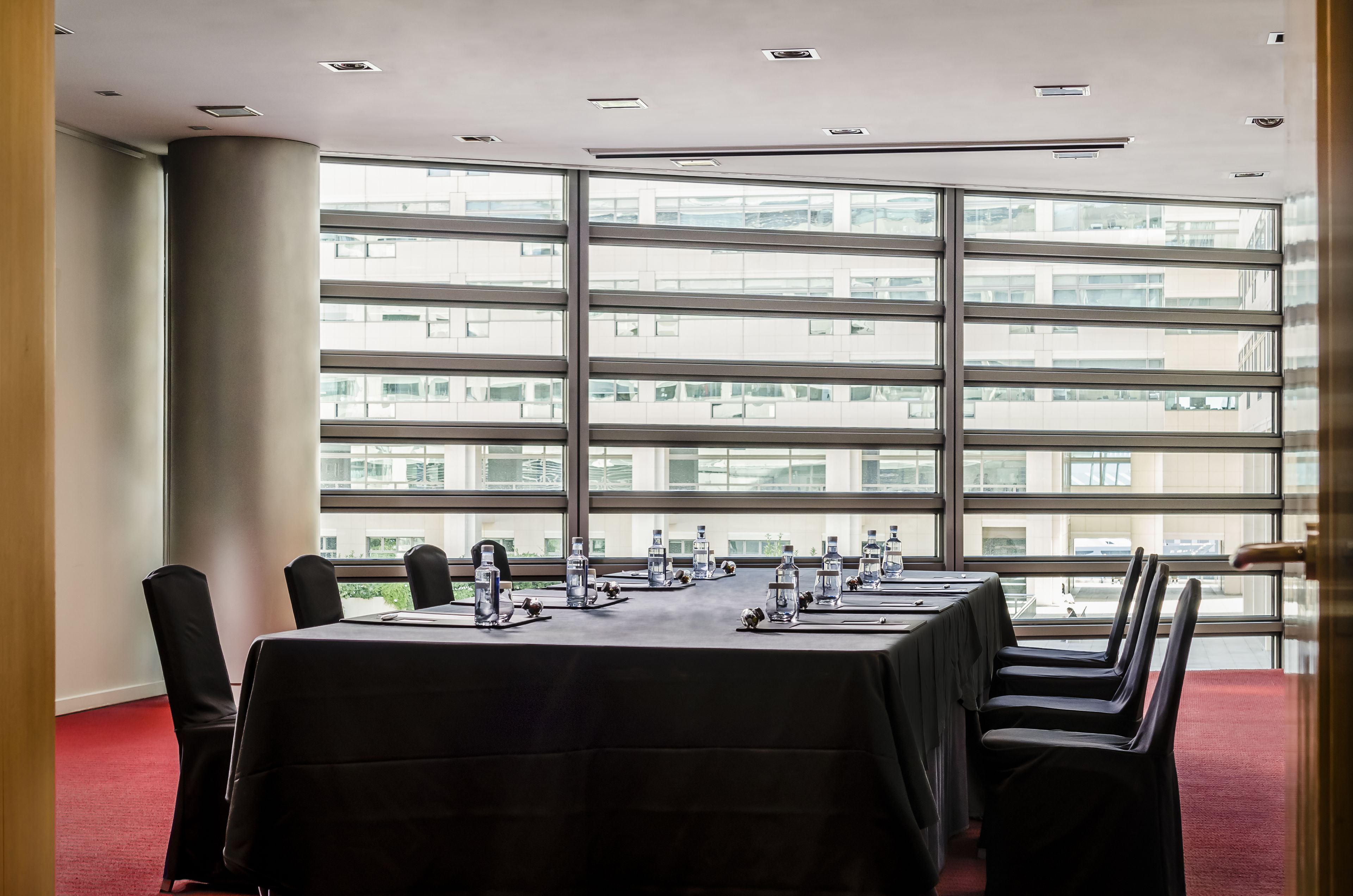 A conference room with a long table, chairs, water bottles, and a view of a high-rise building through large windows.