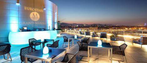 A rooftop terrace with tables and chairs, a pool, and a view of the cityscape at dusk.