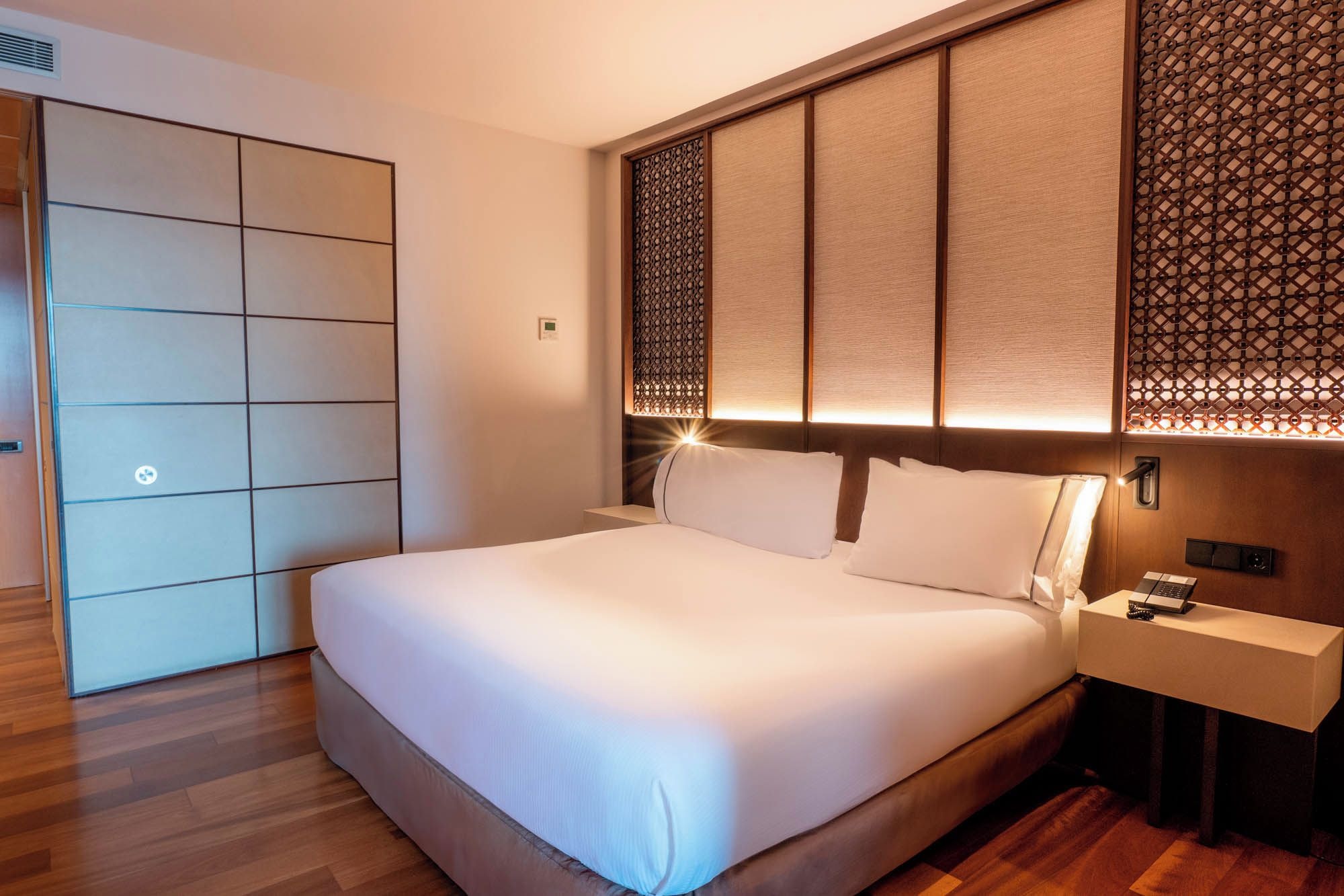 A hotel room with a large bed, a bedside table, a wardrobe, and a decorative wall panel.