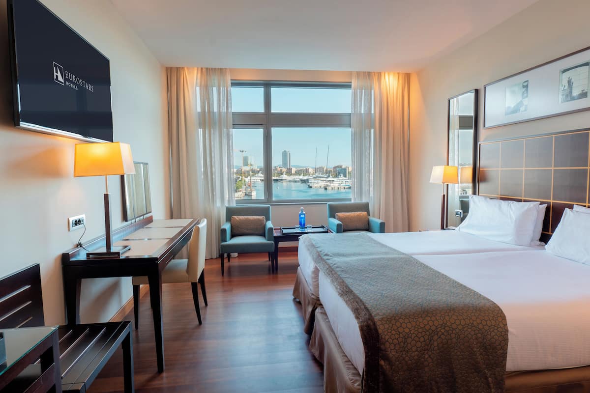Een hotelkamer met een groot bed, een bureau, twee stoelen, een televisie en uitzicht op de jachthaven.