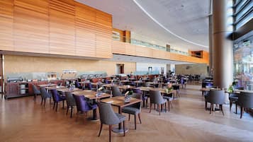 A spacious, modern dining area with multiple tables and chairs, a kitchen in the background, and a high ceiling with a unique design.