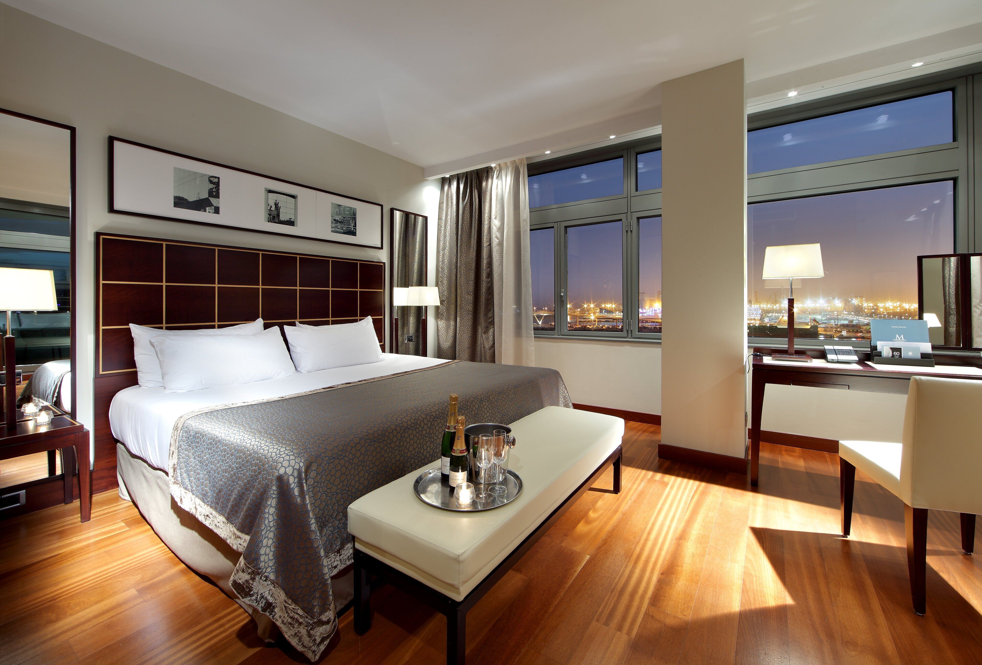 Habitación de hotel con una cama grande, un escritorio con lámpara, vista de la ciudad de noche y una bandeja con champán y copas.