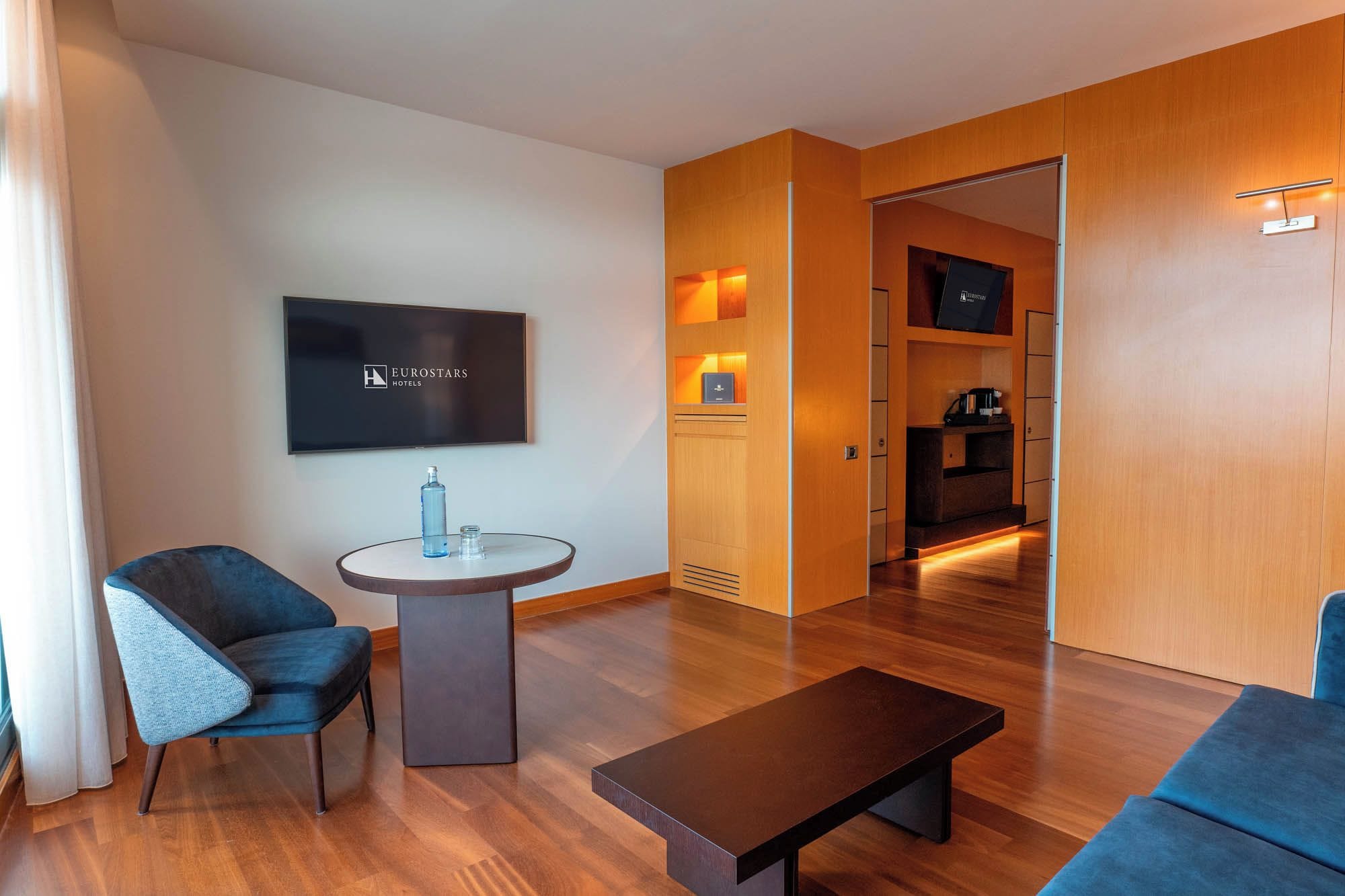 Una habitación de hotel moderna con un televisor de pantalla plana, una mesa redonda con una botella y un vaso, un sillón azul, un banco de madera oscura y un armario de madera.