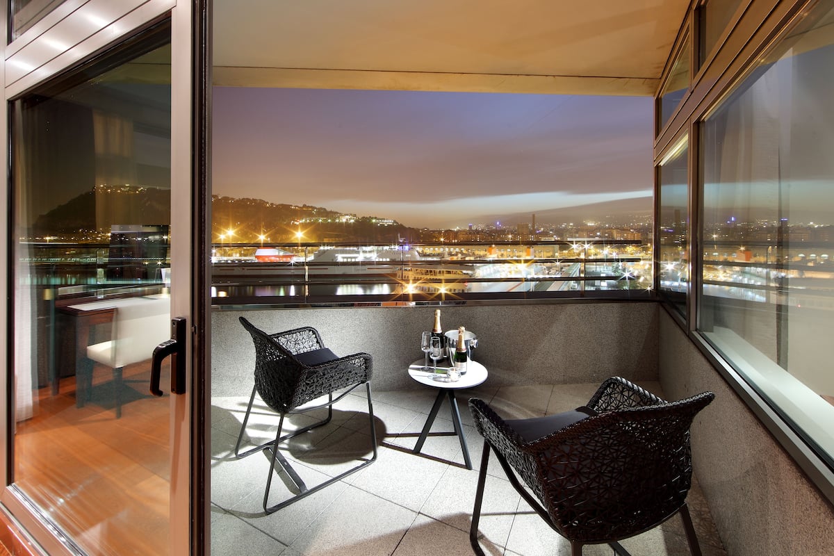 Een balkon met twee rieten stoelen, een klein rond tafeltje met een fles en glazen, en een stadszicht in de schemering.