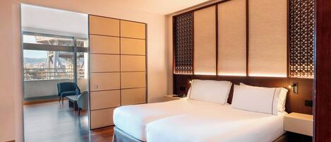 A hotel room with a large bed, a desk, and a view of a cityscape through a large window.