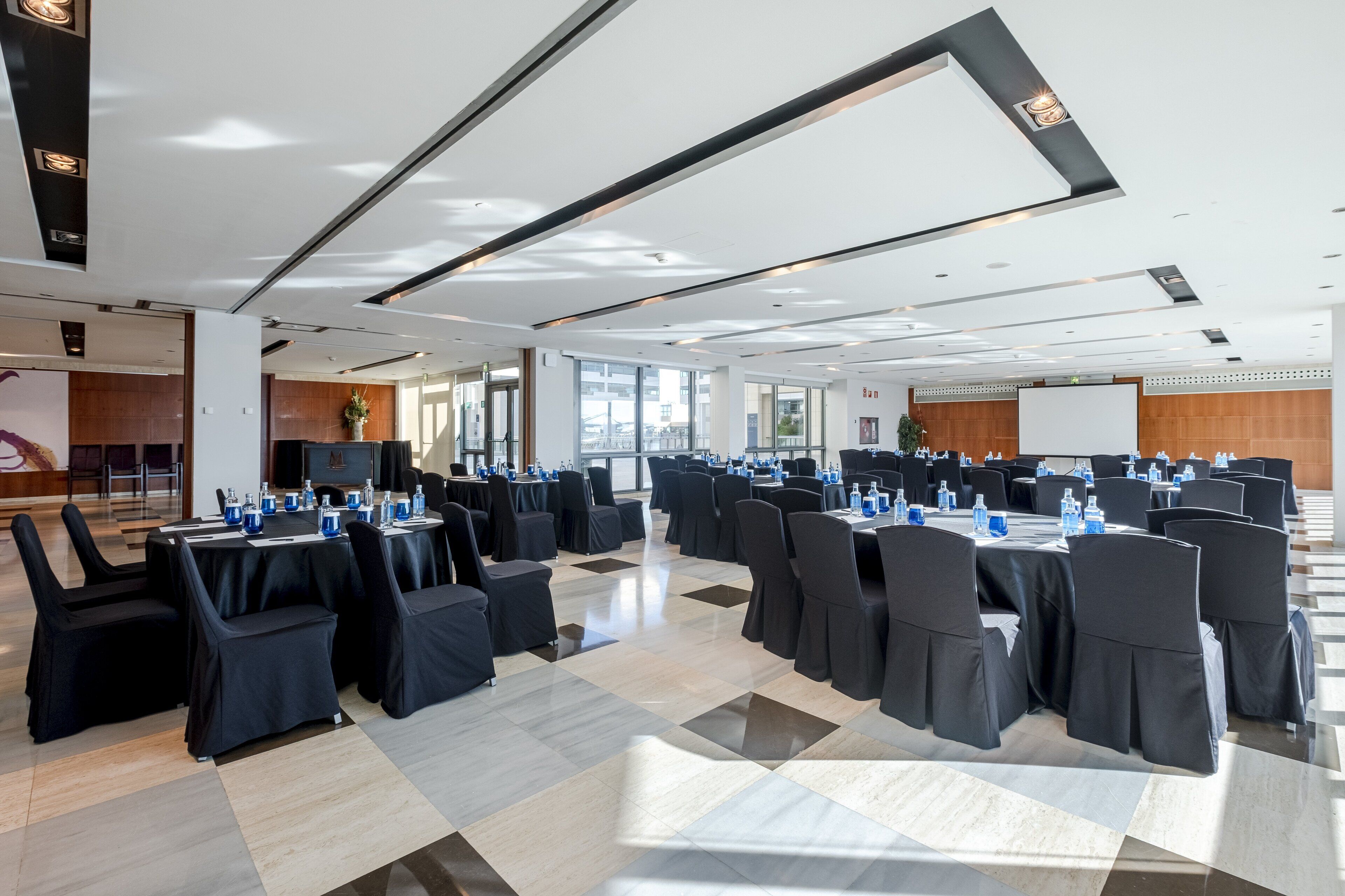 A large conference room with multiple tables and chairs, equipped with water bottles and nameplates, in a modern building with large windows and a high ceiling.