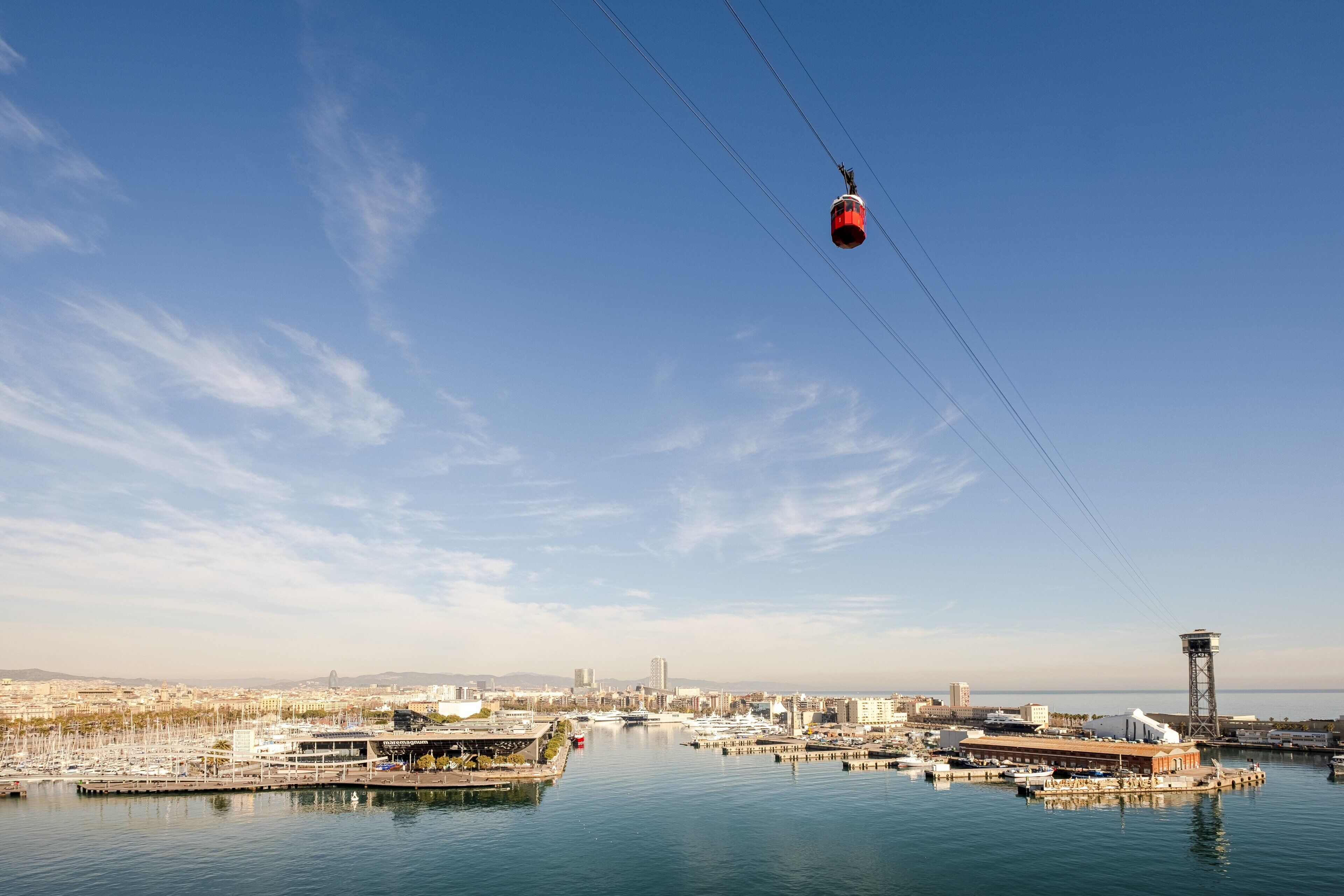 Un teleférico se desplaza sobre una masa de agua, con un puerto deportivo y edificios al fondo.