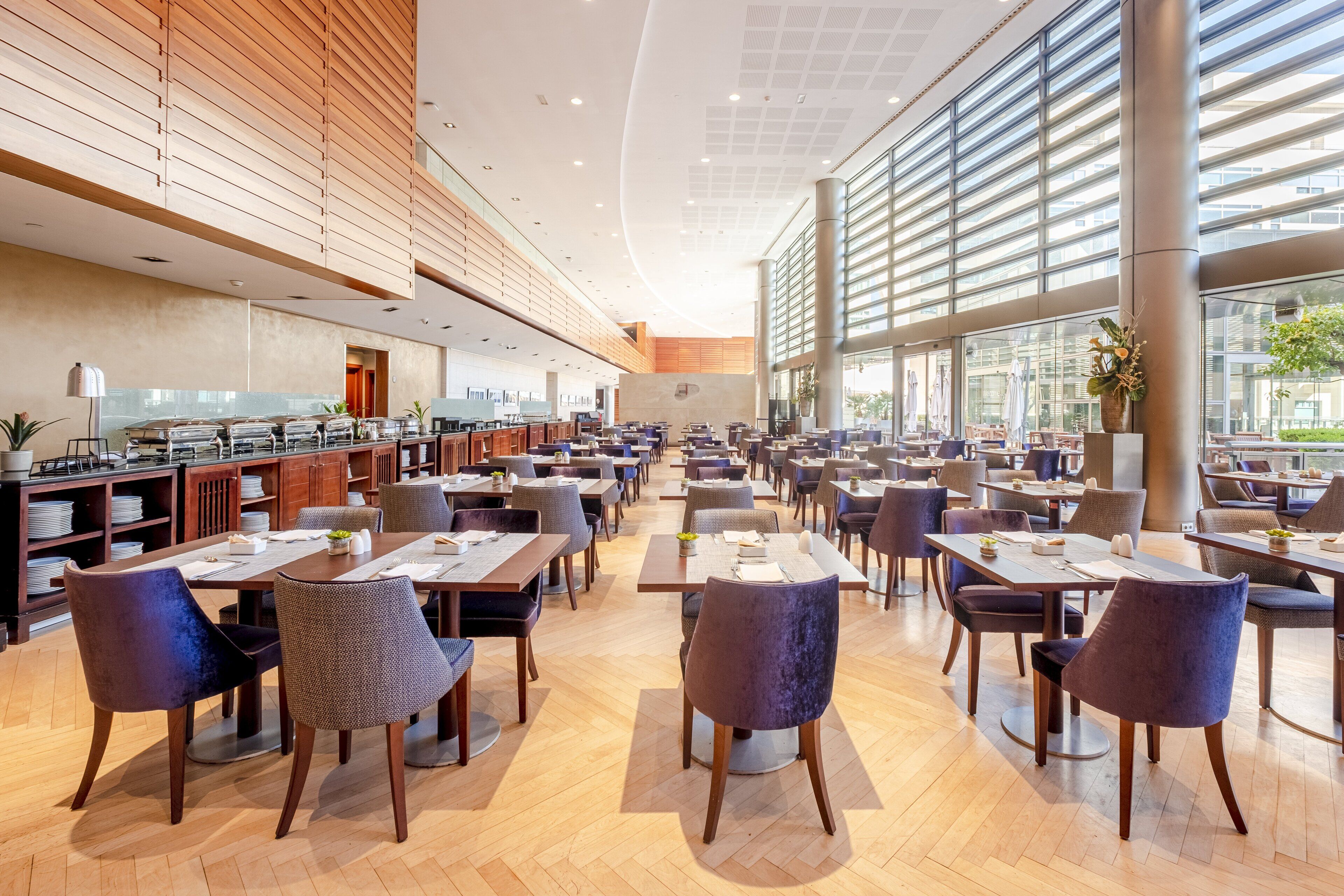 Un grand espace dédié à la salle à manger, avec plusieurs tables et chaises, un coin cuisine doté d’étagères et de grandes fenêtres qui laissent entrer la lumière naturelle.
