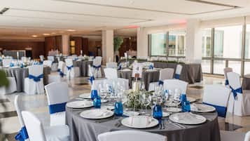 A banquet hall with round tables set for a formal event, featuring white chairs with blue sashes, dark tablecloths, and blue glassware.
