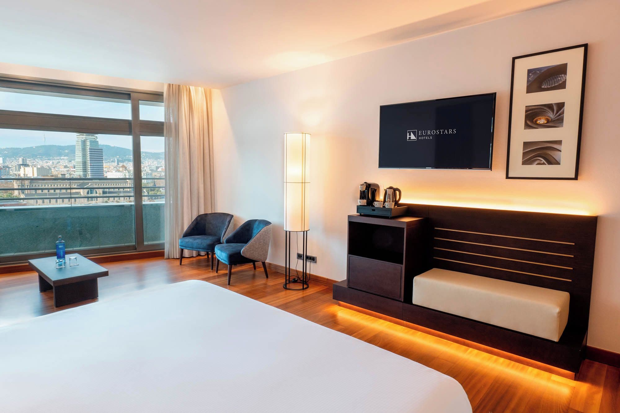 Habitación de hotel con una cama grande, un escritorio, una silla, un televisor y vistas a la ciudad.