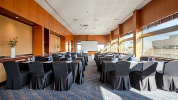 Un salón de conferencias con filas de mesas y sillas, una pantalla grande y vistas al exterior a través de amplias ventanas.