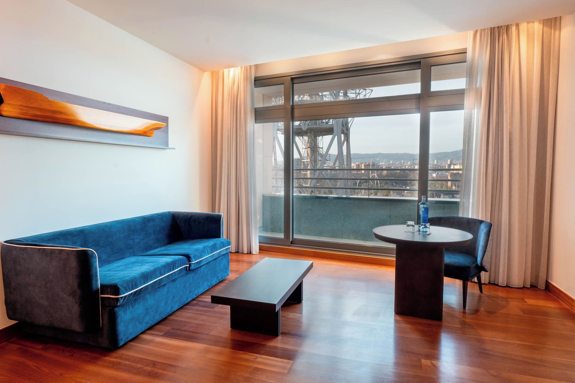Een moderne woonkamer met een blauwe bank, een klein tafeltje en een stoel. Een groot raam biedt uitzicht op een brug en stadsbeeld.