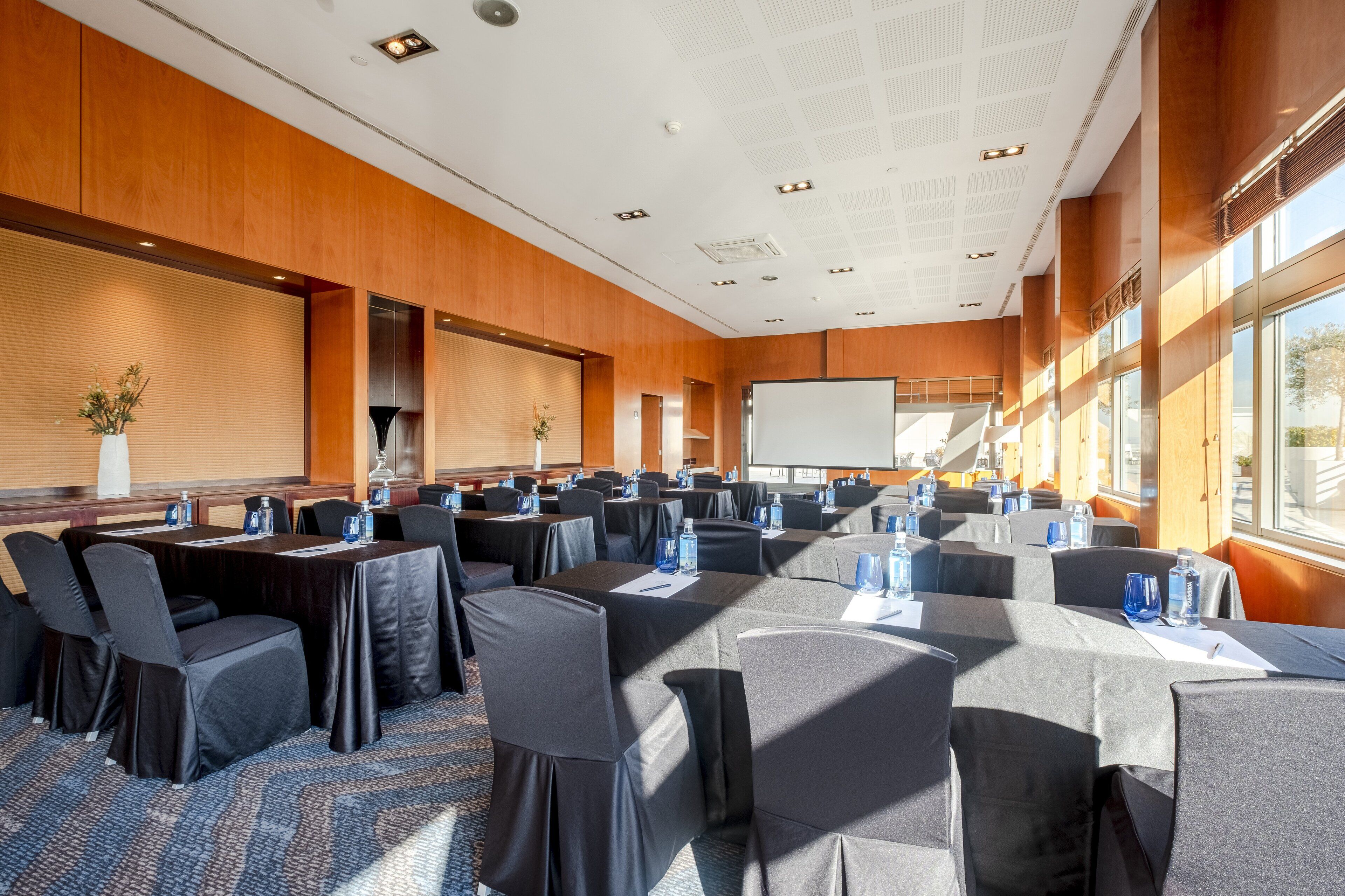 Un salón de conferencias con varias mesas y sillas, equipado con botellas de agua y tarjetas de identificación, ventanas amplias y una pizarra.