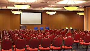 Une salle de conférence avec des rangées de chaises rouges, un grand écran et un éclairage au plafond.