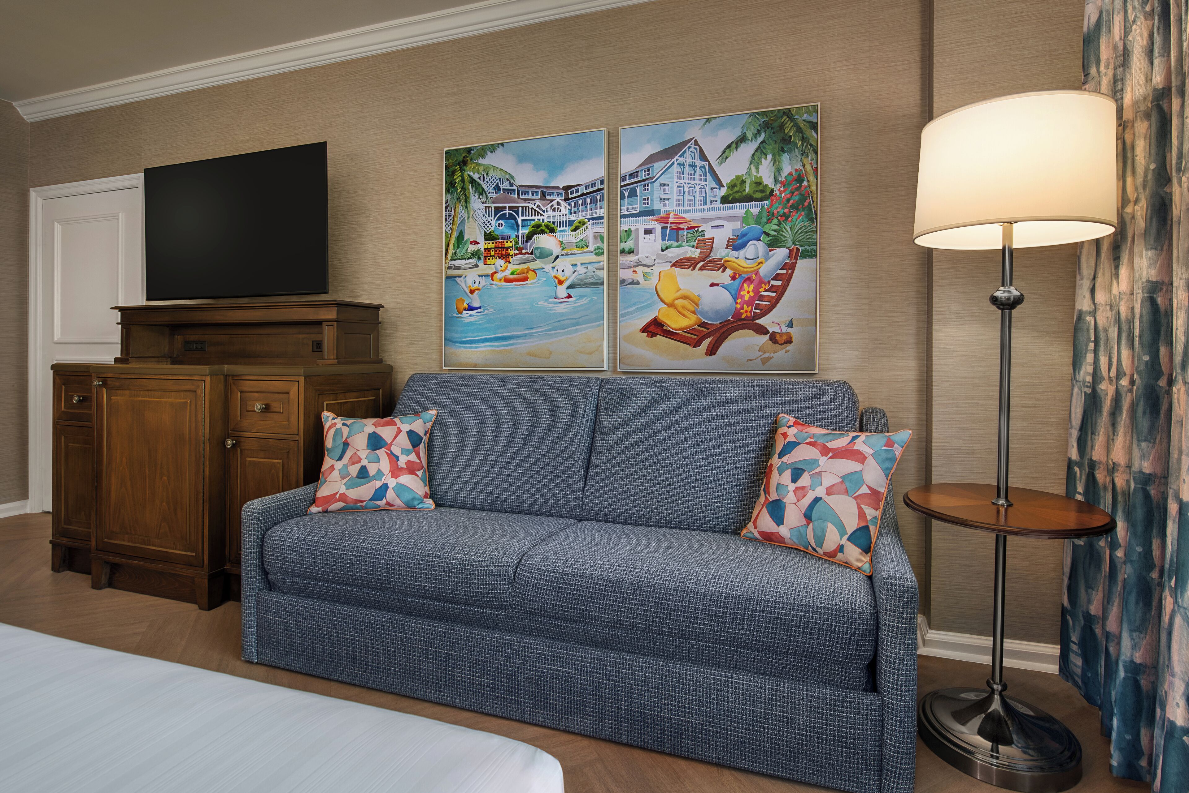 A hotel room with a large sofa, two patterned throw pillows, a flat-screen TV on a wooden cabinet, and two wall art pieces depicting tropical scenes.