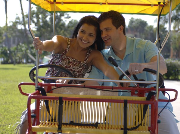 A golf cart with two people, one driving and one sitting, on a grassy area with trees in the background. - Disney's Beach Club Resort (Lake Buena Vista)