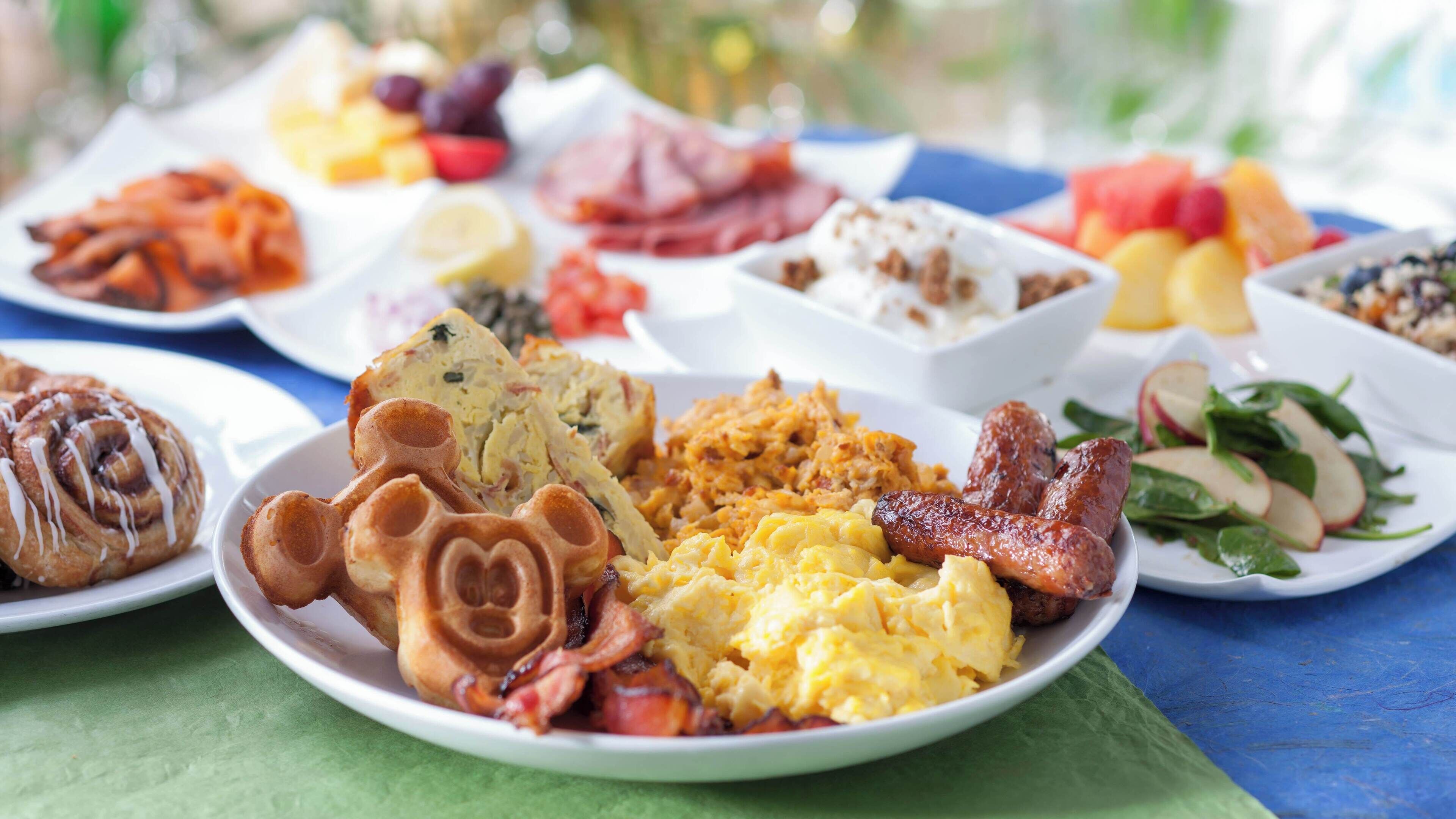 A breakfast buffet with various dishes including waffles, scrambled eggs, sausages, and fresh fruits.