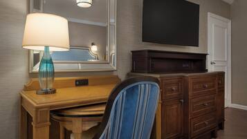 Une chambre d’hôtel avec un bureau en bois, une chaise à rayures bleues, une lampe, une commode et un téléviseur à écran plat fixé au mur.