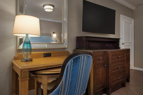 A hotel room with a wooden desk, a blue-striped chair, a lamp, a dresser, and a flat-screen TV mounted on the wall. - Disney's Beach Club Resort (Lake Buena Vista)
