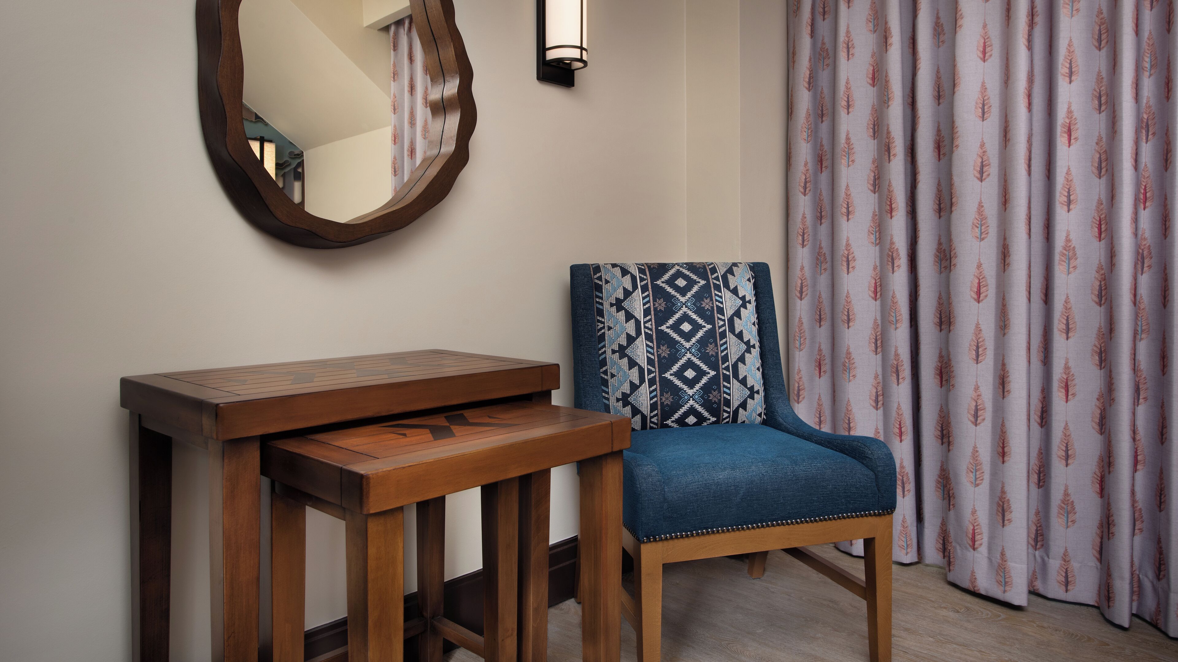 A room with a wooden table, a blue upholstered chair with a patterned cushion, a wall-mounted mirror, and a curtain with a leaf pattern.