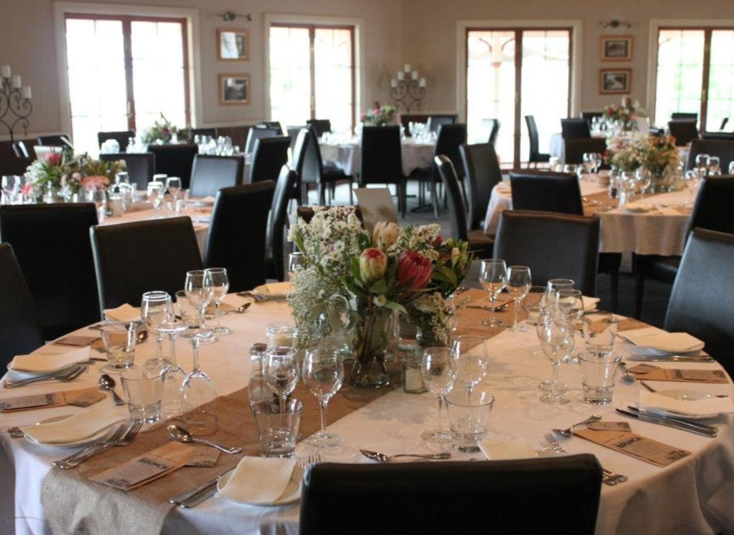 Ein Essbereich mit runden Tischen, gedeckt für eine Mahlzeit, wobei jeder Tisch eine Blumenarrangemement und schlichte Gläser ziert.