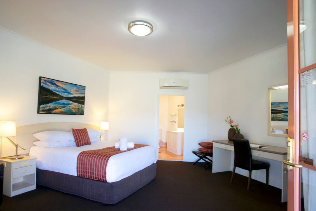 A hotel room with a bed, bedside tables, a desk, a chair, a mirror, and a painting on the wall.
