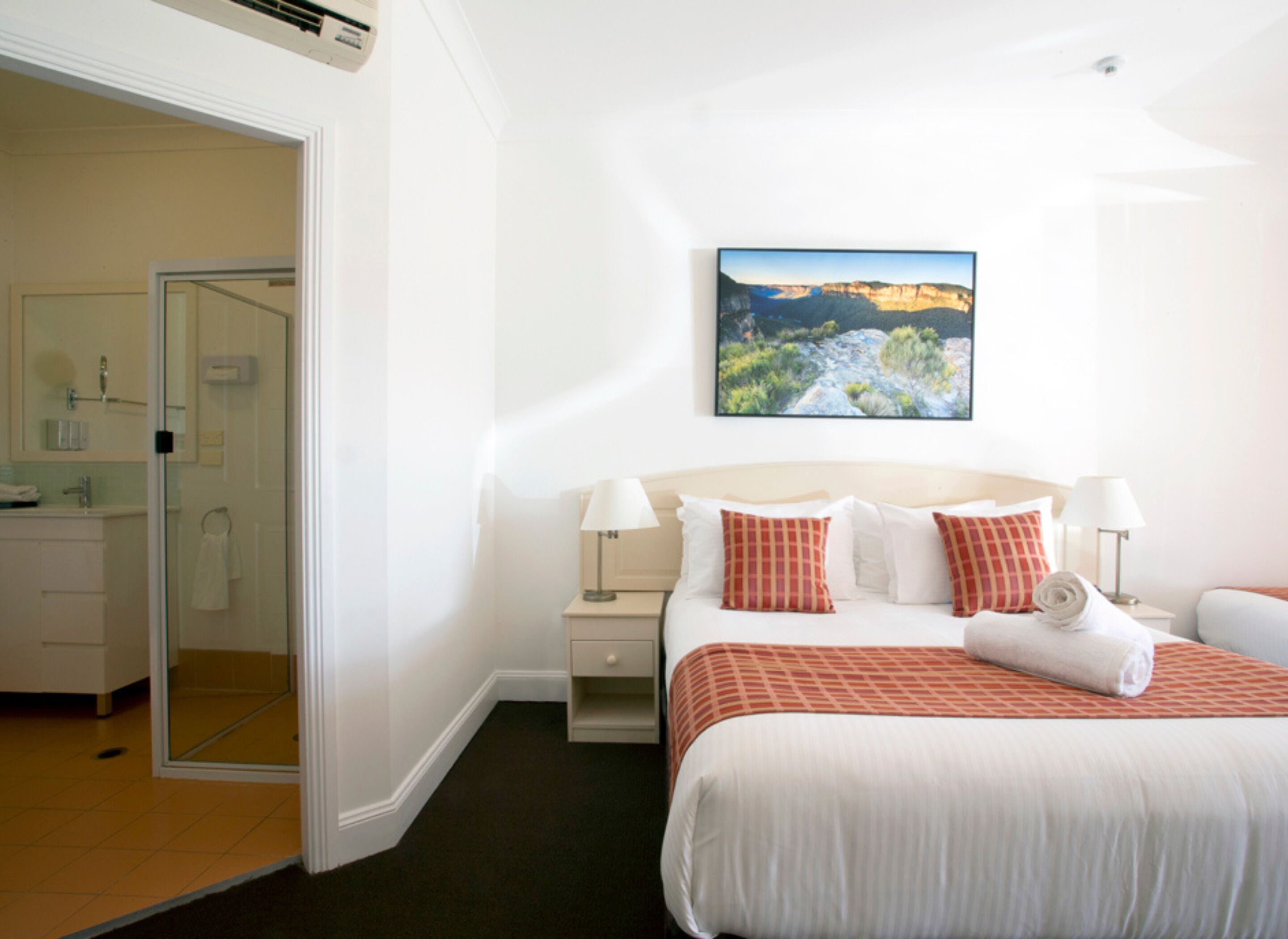 A hotel room with a bed, a nightstand, a painting, and a bathroom visible through an open door.