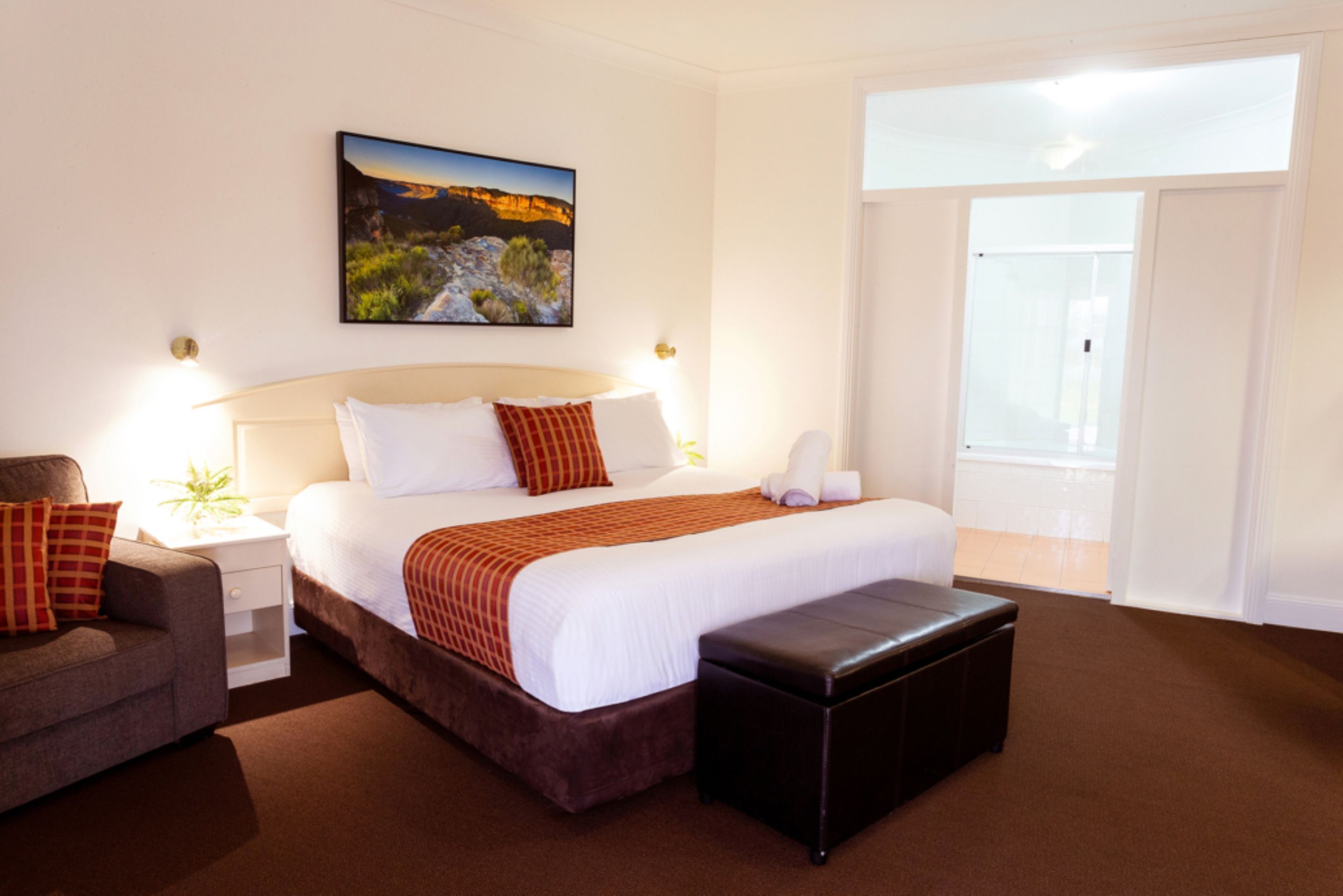 A hotel room with a large bed, a nightstand, a chair, a bench, and a painting on the wall.