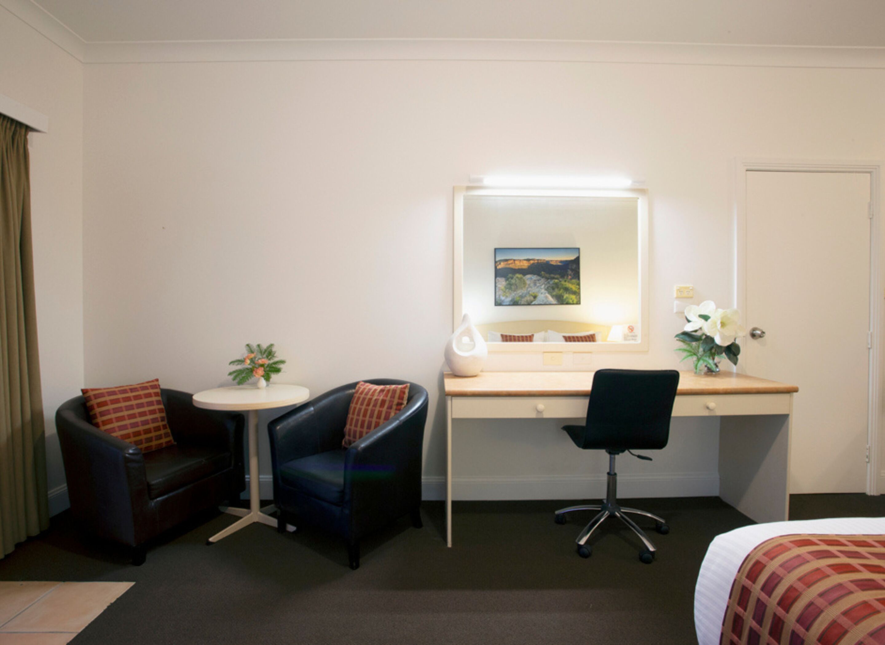Habitación de hotel con cama, escritorio con espejo, dos sillones, una mesita redonda con una planta y una puerta.