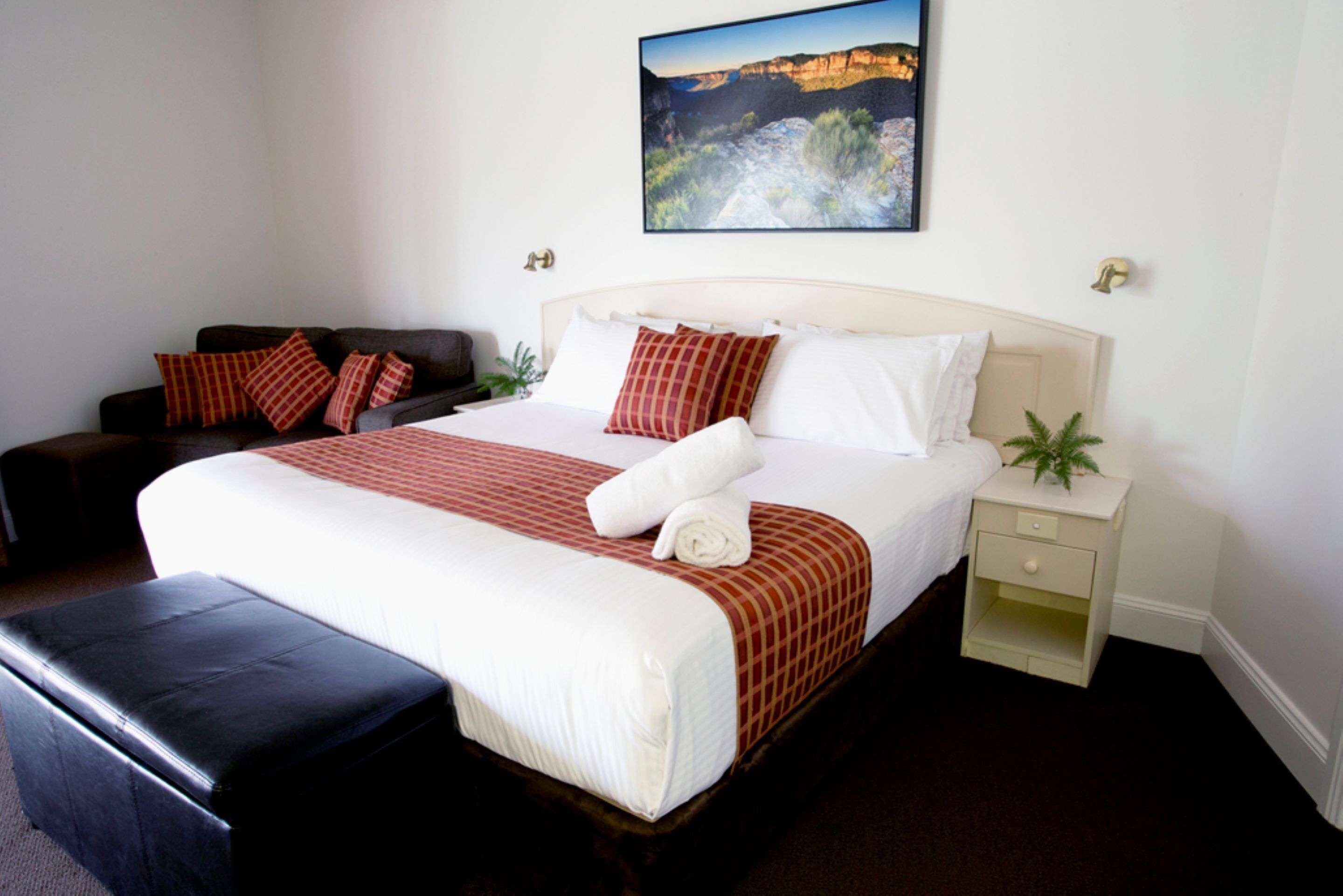 Una habitación de hotel ordenada con una cama grande, un sofá, una mesita de noche y un cuadro colgado en la pared.