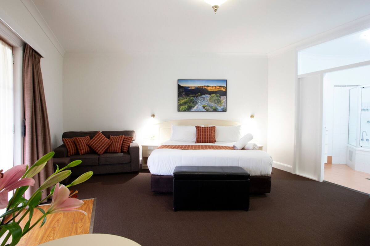 A hotel room with a bed, a sofa, a nightstand, a lamp, a painting on the wall, and a window with curtains.