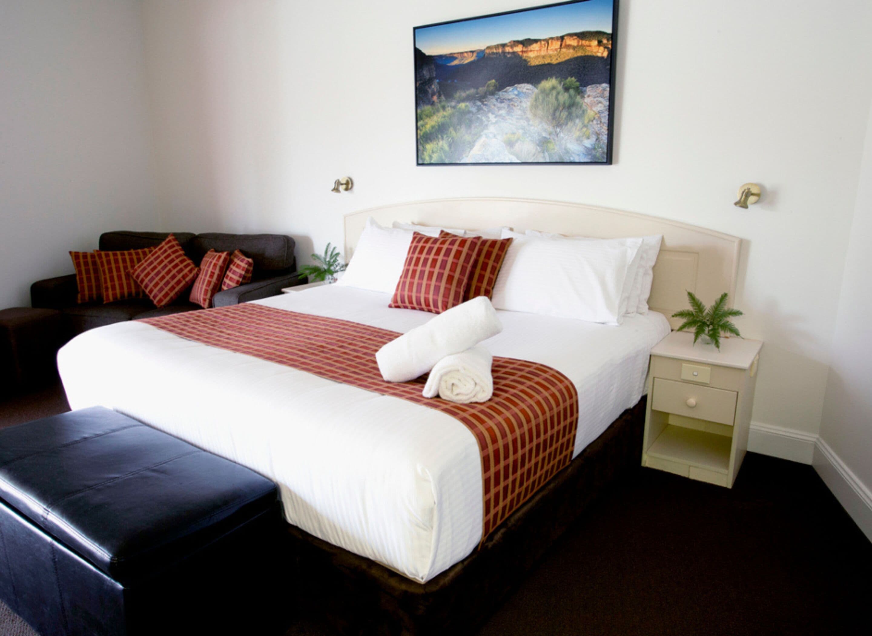 A neatly made bed with a red and white checkered bedspread, a white pillow, and a rolled white towel. There is a dark brown sofa with red and white pillows to the left. A small green plant is on the nightstand beside the bed.