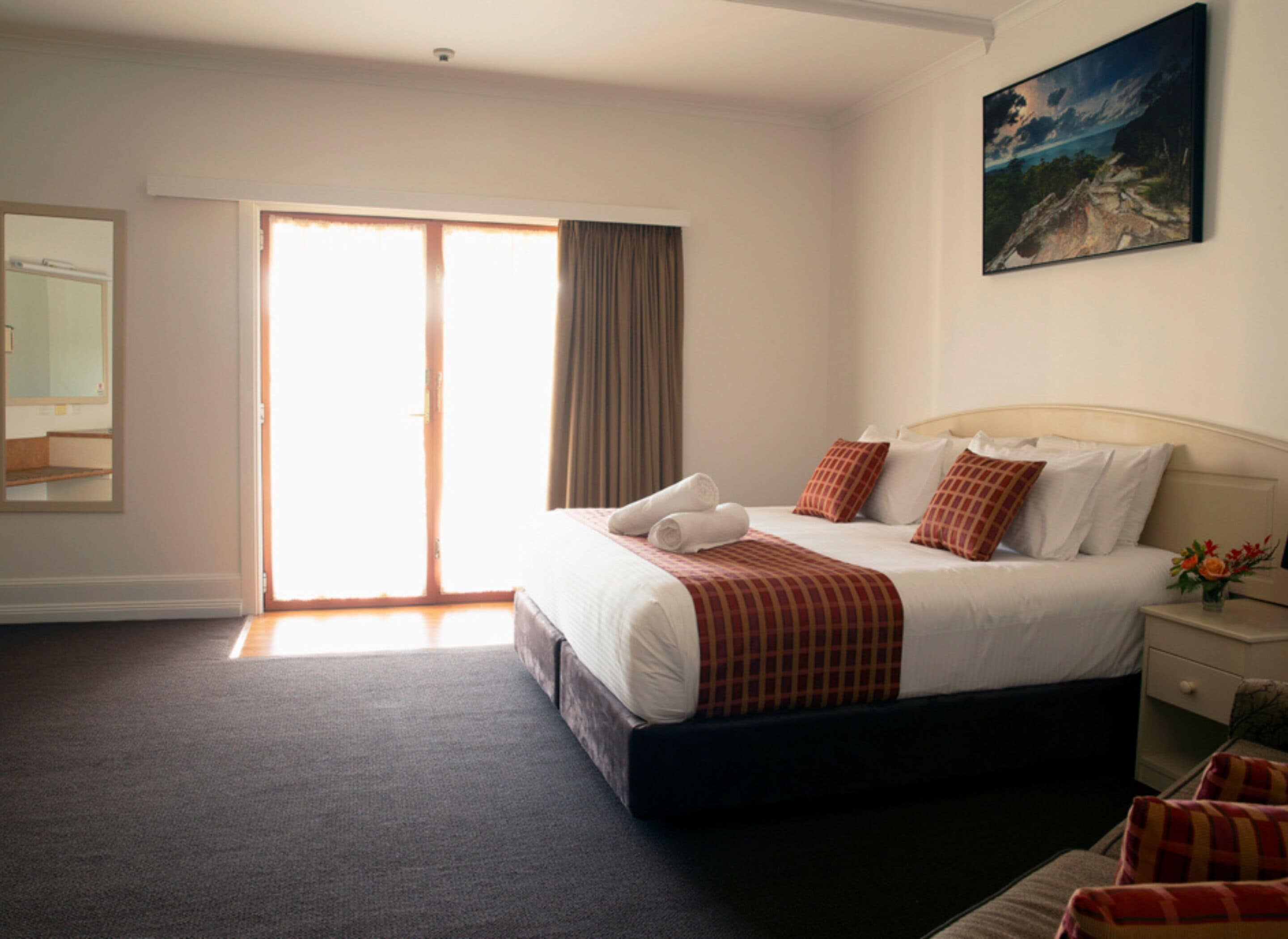A hotel room with a large bed, a painting on the wall, a mirror, and a sliding door to the outside.